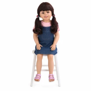 Life-like doll with brown hair in pigtails, wearing a pink shirt and denim dress, seated on a white chair.